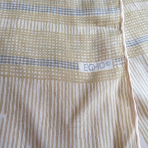 May include: A lightweight scarf featuring a pattern of vertical tan stripes and horizontal bands of tan and light blue grid lines. The fabric has a soft texture with a finished edge and includes the brand name ECHO printed in a white rectangular box.