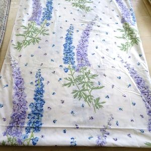 Puede incluir: Tela blanca con un patrón repetido de flores azules y moradas. Las flores están dispuestas en líneas verticales y están rodeadas de pequeñas flores azules dispersas.