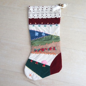 Puede incluir: Un calcetín navideño decorativo con un diseño de patchwork de paneles de terciopelo y tela en tonos burdeos, crema, azul, verde oliva y verde oscuro. El puño tiene encaje blanco y una cinta para colgar, con detalles de bordados florales.