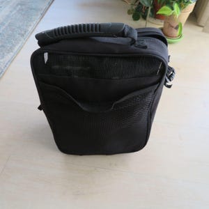 May include: A black camera bag with a handle on top and a mesh pocket on the front. The bag has a rectangular shape and a side pocket. The bag is sitting on a light-colored wooden floor.