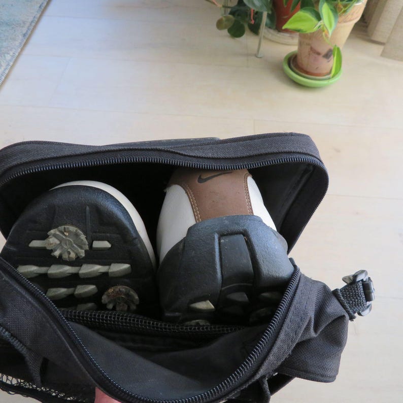 May include: Black zippered golf shoe bag with two golf shoes inside. The shoes are white, brown, and black with the Nike logo. The bag is open, revealing the soles of the shoes.