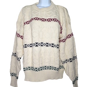 Puede incluir: Un suéter de punto color crema con un patrón de punto de cable y rayas de colores. Las rayas son rojas, azules y verdes.