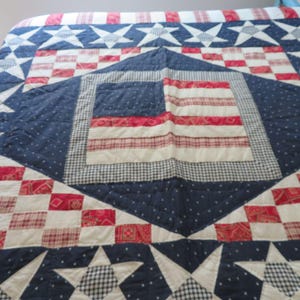 May include: A patchwork quilt featuring a central American flag design surrounded by a navy blue field with white stars. The quilt incorporates red, white, and blue fabric squares in various patterns including plaid, gingham, and floral prints.