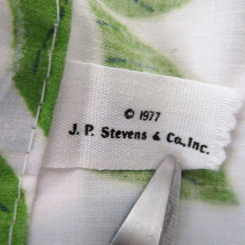 Puede incluir: Primer plano de una etiqueta de tela blanca con texto negro: "&copy; 1977 J.P. Stevens & Co., Inc." La etiqueta se est&aacute; cortando con unas tijeras. El fondo muestra tela blanca con estampados de hojas verdes.