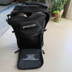 May include: Black nylon bag with a handle and multiple compartments. The bag has a rectangular patch with the letters "PCG" in white. The bottom compartment is open, revealing text in white. The bag is on a light-colored floor.