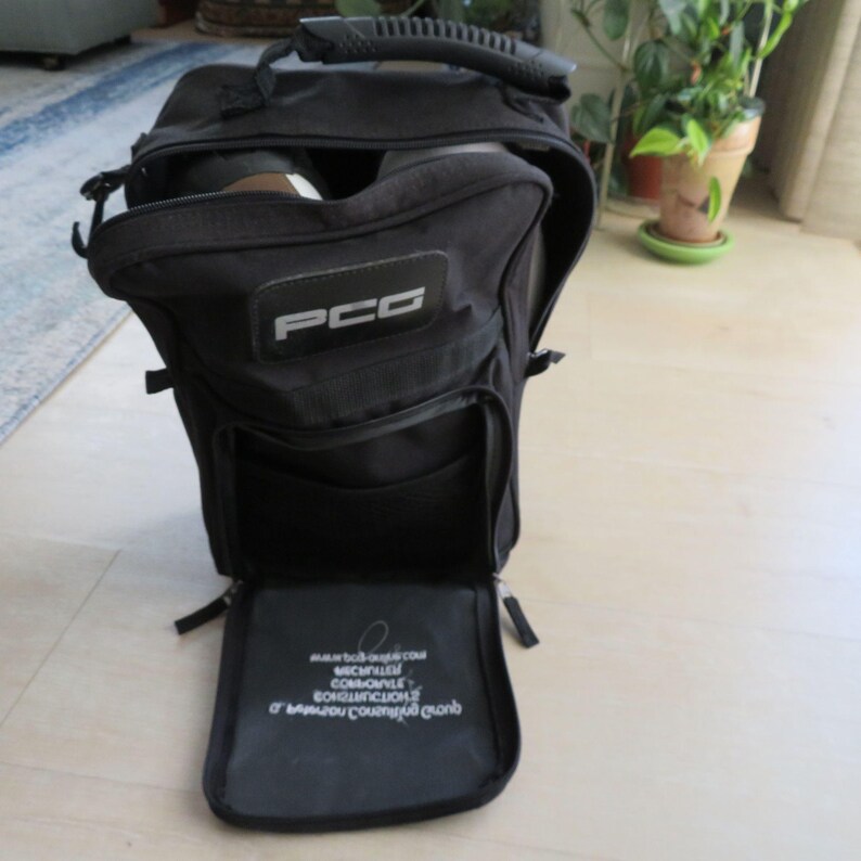 May include: Black nylon bag with a handle and multiple compartments. The bag has a rectangular patch with the letters "PCG" in white. The bottom compartment is open, revealing text in white. The bag is on a light-colored floor.