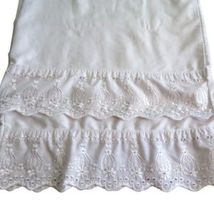 Puede incluir: Tela blanca de ojal con borde festoneado. La tela tiene un patrón detallado de flores y hojas. El diseño de ojal se repite en dos filas, creando un efecto en capas. La tela probablemente se utiliza para ropa o decoración del hogar.