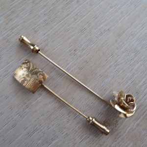 Può includere: Due spille da bavero color oro. Una spilla presenta una testa rettangolare con un disegno inciso, e l'altra ha una testa a forma di rosa. Entrambe le spille hanno gambi lunghi e sottili e estremità decorative.