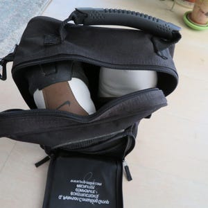 May include: Black sports bag with a top handle and a zippered main compartment. Inside, a pair of athletic shoes are visible. One shoe is brown, white, and black with a Nike logo. The bag has a small zippered pocket on the front.