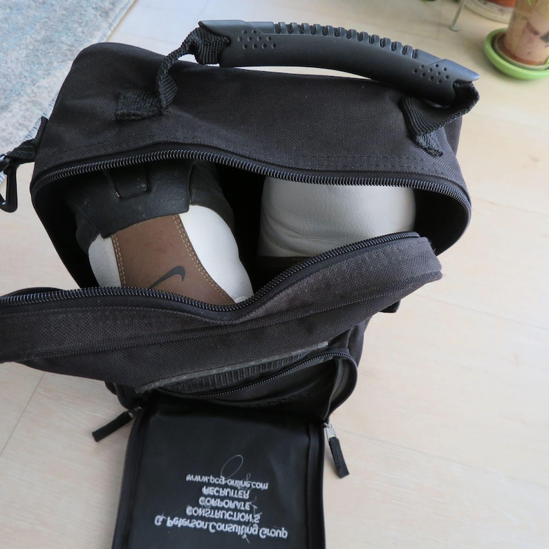 May include: Black sports bag with a top handle and a zippered main compartment. Inside, a pair of athletic shoes are visible. One shoe is brown, white, and black with a Nike logo. The bag has a small zippered pocket on the front.