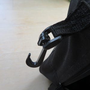 May include: Close-up of a black plastic hook attached to a black fabric strap. The hook is curved and appears to be part of a bag or carrying case, designed for securing the strap.