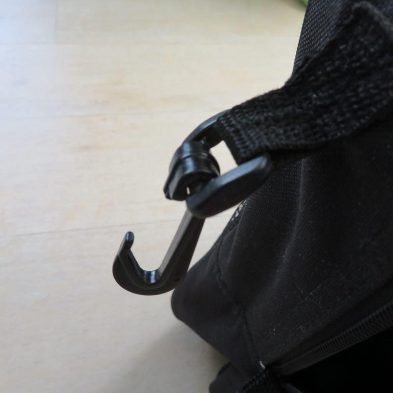 May include: Close-up of a black plastic hook attached to a black fabric strap. The hook is curved and appears to be part of a bag or carrying case, designed for securing the strap.