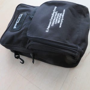 May include: Black canvas bag with multiple compartments and a shoulder strap. The bag features the text "PCG" on a patch and the text "G. Peterson Consulting Group CONSTRUCTION'S CORPORATE RECRUITER www.pcg-online.com" printed in white.
