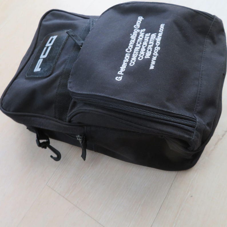 May include: Black canvas bag with multiple compartments and a shoulder strap. The bag features the text "PCG" on a patch and the text "G. Peterson Consulting Group CONSTRUCTION'S CORPORATE RECRUITER www.pcg-online.com" printed in white.