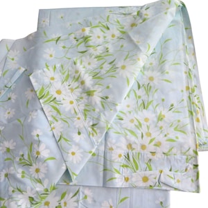 Puede incluir: Juego de sábanas azul claro con un estampado floral de margaritas. Las sábanas presentan margaritas blancas con centros amarillos y tallos y hojas verdes. El diseño floral se extiende por las sábanas.