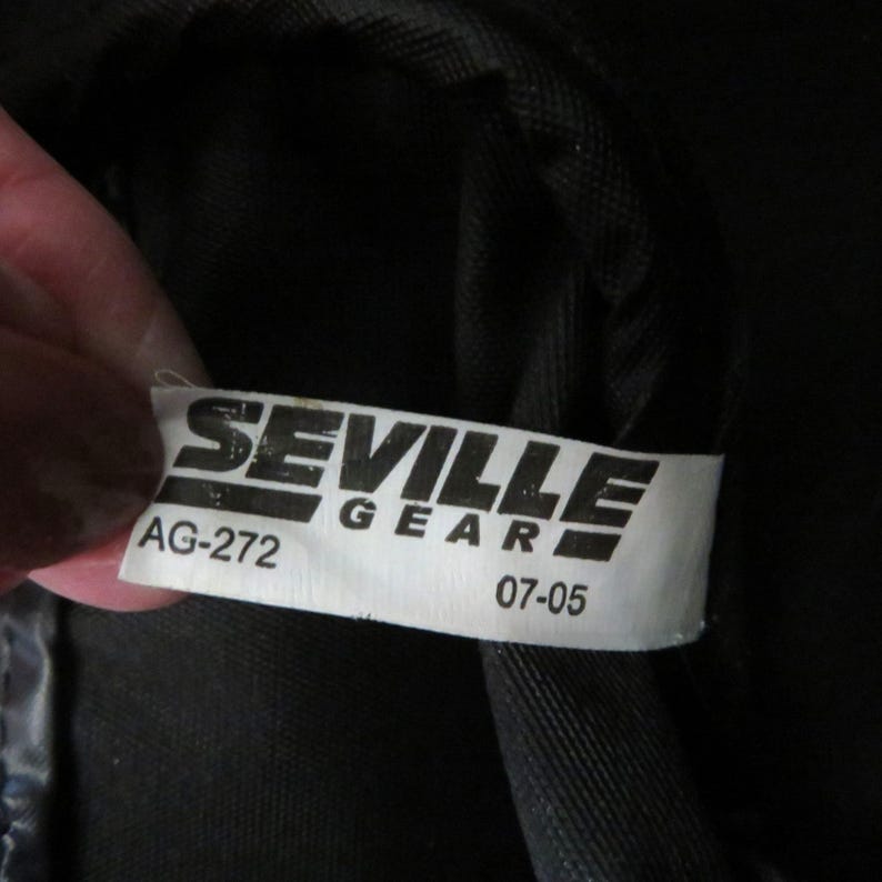 May include: A close-up of a white tag with the words "SEVILLE GEAR" printed in black. Below the text are the numbers "AG-272" and "07-05". The tag is attached to a black fabric.