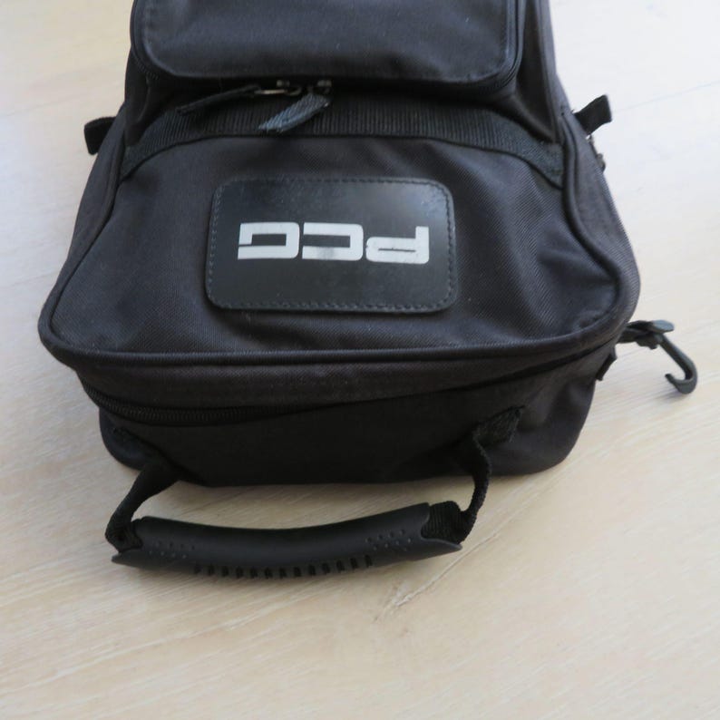 May include: Black nylon bag with a top handle and a zippered main compartment. The bag features a rectangular patch with the letters "PCG" in white. A smaller zippered compartment is visible on the front.