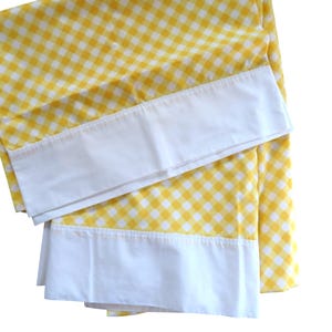 Peut inclure: Une taie d'oreiller à motif vichy jaune et blanc dotée d'une large bordure en tissu blanc uni le long de l'ouverture. Le textile présente un motif à carreaux classique avec un aspect souple et plié, soulignant le contraste entre le motif jaune vif et l'ourlet blanc net.