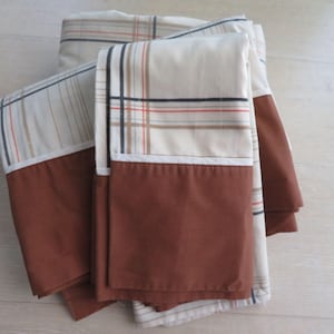 May include: Folded bedding set featuring a cream-colored fabric with a plaid pattern in brown, black, and red. The bottom portion of each piece is a solid brown color. The set appears to be made of a soft, possibly cotton, material.