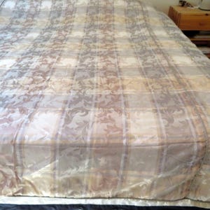 May include: A bedspread featuring a plaid pattern in shades of cream, beige, and muted brown. The fabric displays an intricate, repeating damask-style floral motif woven throughout the checkered design, finished with a decorative piped edge.