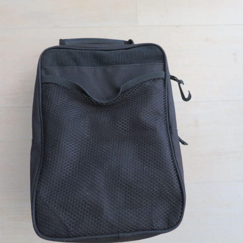 May include: A black travel bag with a mesh pocket on the front. The bag has a handle on top and a hook on the side. The bag is rectangular and appears to be made of a durable material. The bag is designed for carrying items.