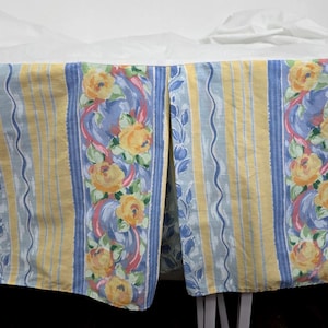 May include: A bed skirt with a floral pattern. The skirt features vertical stripes in yellow and blue, with a central panel of yellow roses, blue ribbons, and green leaves. The fabric is visible against a white background.
