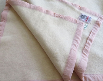 Puede incluir: Una manta de lana blanca con ribete de satén rosa. La manta tiene una etiqueta que dice "The Wool Bureau, Inc. Certified Wool Product."