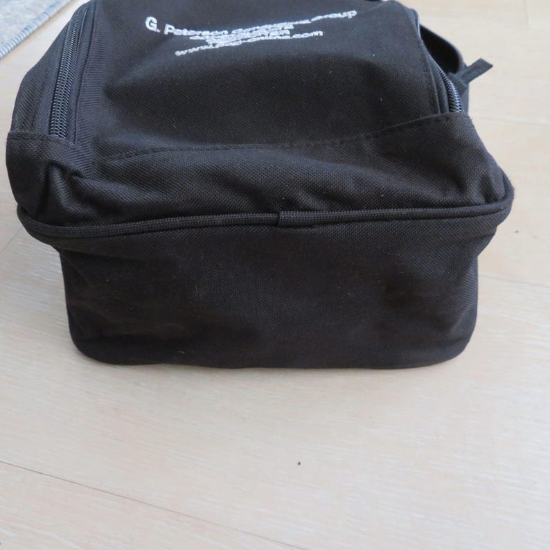 May include: A black rectangular bag with a zipper closure. The bag has a structured shape and appears to be made of a durable fabric. White text is printed on the top of the bag.
