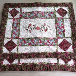 Op de afbeelding: Een gewatteerde deken met een bloemenpatroon. De deken heeft een wit midden met een geborduurd bloemenpatroon, omgeven door panelen van bloemenstof in rood, blauw en groen. De rand is een donkerbruine bloemenprint.