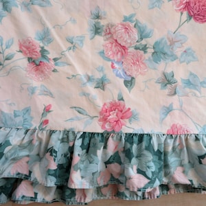 Puede incluir: Un tejido textil con fondo color melocotón claro y un estampado floral de rosas rosas y hojas verde salvia. El borde inferior está rematado con un volante fruncido en un tono verde bosque más oscuro con un estampado floral a juego.