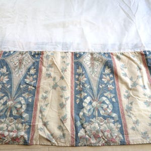 May include: A white bed skirt with a floral print in shades of blue, green, and pink. The skirt has a ruffled edge and is made of a soft, cotton fabric.