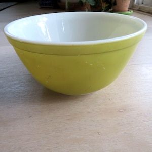 May include: A vintage, lime green mixing bowl with a white interior. The bowl has a rounded shape and a slightly flared rim. The exterior is a solid, muted green color, while the interior is bright white. A thin, gold-colored band encircles the bowl near the rim.