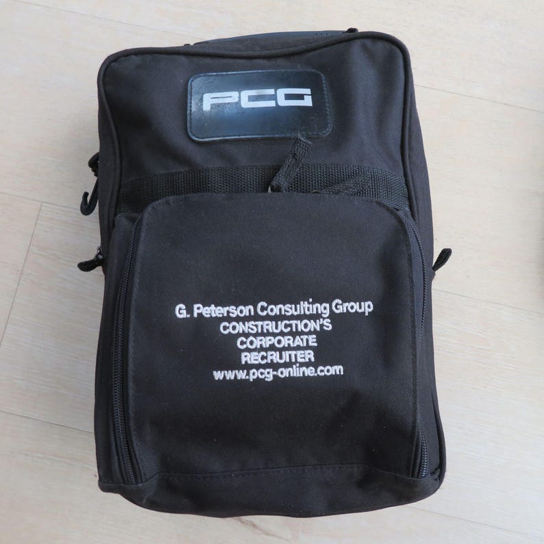 May include: Black rectangular bag with a top handle and a front pocket. The bag has a white logo that says "PCG" on the top and the text "G. Peterson Consulting Group CONSTRUCTION'S CORPORATE RECRUITER www.pcg-online.com" on the front.