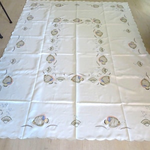 Peut inclure: Une nappe blanche à bord festonné, ornée d'un motif répété de poissons et de coquillages stylisés dans des tons de gris, de bleu et de rose. Le motif est disposé en bordure et en carré central.