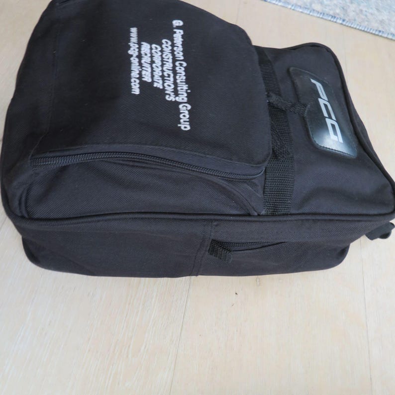May include: A black rectangular bag with a handle and a zippered compartment. The bag has the text "G. Peterson Consulting Group CONSTRUCTION'S CORPORATE RECRUITER www.pcg-online.com" printed in white. The bag also has the letters "PCG" on the side.