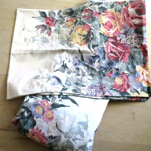 May include: Two white pillowcases with a floral pattern featuring pink, yellow, and red roses, along with blue and purple flowers and green foliage. The bedding has a vintage aesthetic.