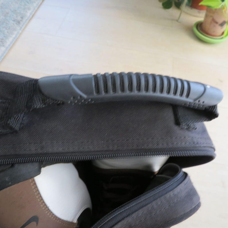 May include: Black sports equipment bag with a textured gray handle. The bag is partially unzipped, revealing white and brown sports equipment. The Nike logo is visible on the brown item.