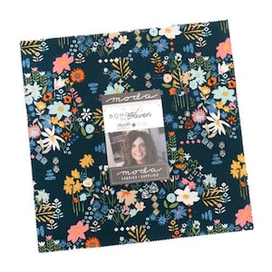 May include: A square fabric piece with a dark teal background, covered in a floral pattern of white, pink, orange, and blue flowers. A label with the text "moda" and "BOH Haven" is attached, featuring a woman's photo.