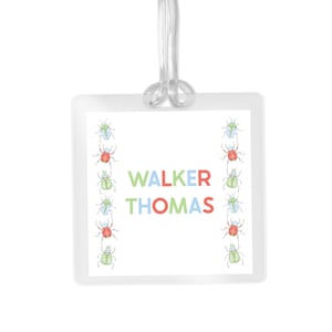 May include: A clear, square luggage tag with the name "WALKER THOMAS" in colorful letters. The tag is decorated with rows of illustrated beetles in green, red, and blue. A clear plastic loop is attached at the top.