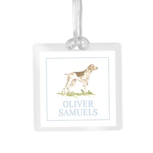 May include: A clear, square luggage tag with a watercolor illustration of a dog. The dog is brown and white, standing on green grass. The text "OLIVER SAMUELS" is printed in light blue below the dog.