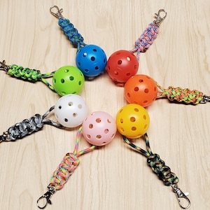 May include: A collection of colorful pickleball keychains. Each keychain features a perforated ball in a different color, including blue, green, red, orange, white, pink, and yellow. The keychains have a braided cord and a metal clasp.