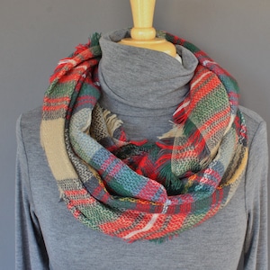 May include: A red, green, and beige plaid infinity scarf with frayed edges. The scarf is worn around the neck of a gray turtleneck sweater.