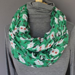 May include: Green infinity scarf with a snowman pattern. The scarf features white snowmen with black hats and red buttons, and red stars on a green background.