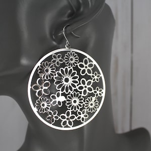 May include: Silver-toned, circular earrings with a floral design. The earrings feature a delicate, openwork pattern of various flower shapes within a circular frame. The earrings are shown on a mannequin head.