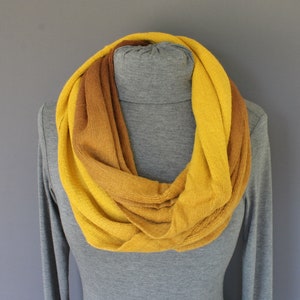 May include: A yellow and brown infinity scarf made of knitted fabric. The scarf is folded in a loop and draped over a gray mannequin.