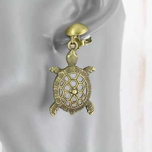 May include: A pair of gold-tone clip-on earrings featuring a detailed turtle design. The turtle's shell is openwork and has a hexagonal pattern.
