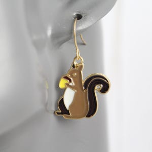 May include: A gold-colored earring featuring a cartoon squirrel design. The squirrel is brown with a white belly, a black tail, and is holding a yellow acorn. The earring is hanging from a gold-colored hook.