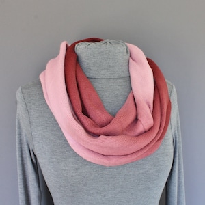 May include: A knitted infinity scarf in shades of pink and red. The scarf is wrapped around a gray turtleneck, showcasing the color gradient from light pink to deep red. The soft texture and color palette suggest a cozy accessory.