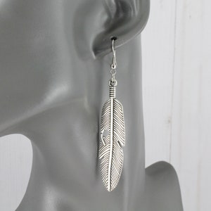 May include: Silver feather dangle earrings. The feather is detailed with lines and has a small hole in the center. The earrings are on a mannequin's ear.