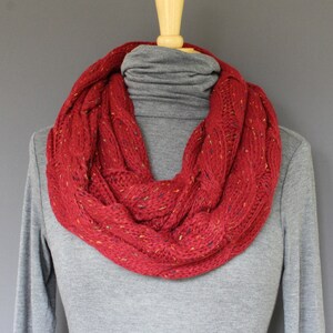 May include: A red infinity scarf with a cable knit design. The scarf is displayed on a mannequin and has flecks of yellow, blue, and orange throughout the knit. The scarf is wrapped around a gray turtleneck.